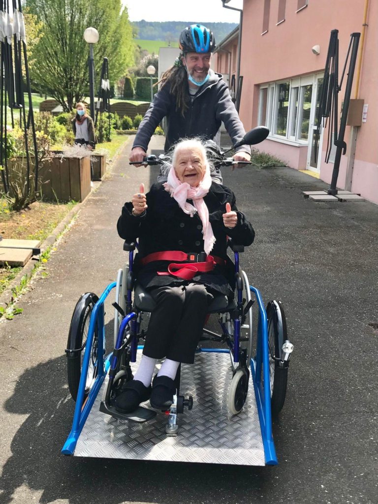 Steve avec Madame G. à l’avant du triporteur pour fauteuil roulant. Elle fait un pouce en l’air, souriante.
