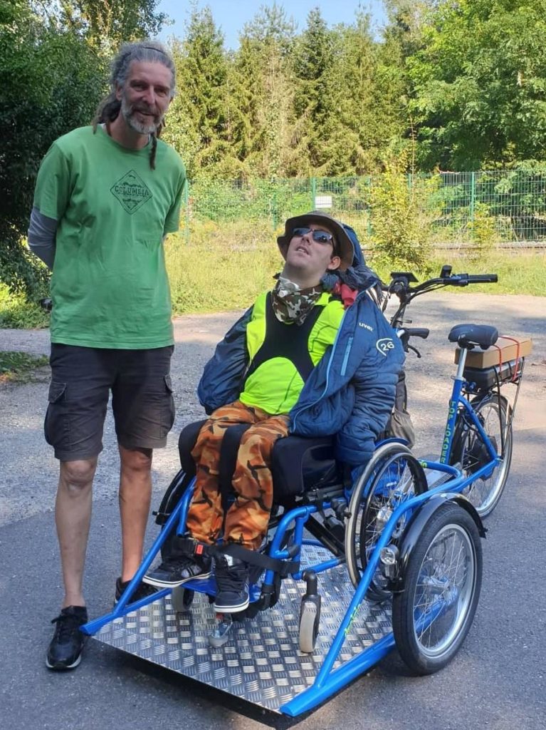 Steve à côté de Mike qui est installé sur un fauteuil roulant à l'avant du triporteur pour PMR.